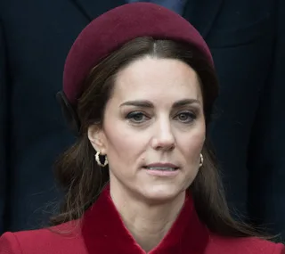 A woman in a red coat and matching hat, with long brown hair and hoop earrings, stands outdoors.