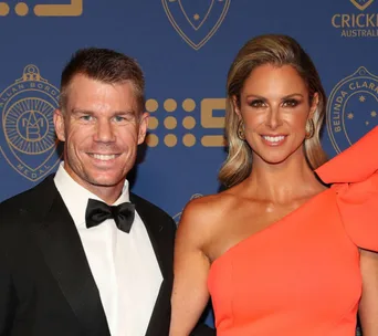 "Man in tuxedo and woman in orange dress smiling at event with Australian cricket logos in background."