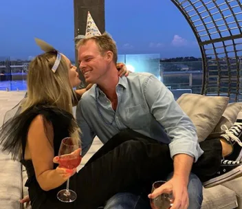 A couple in party hats shares a playful moment, seated closely with wine glasses in hand.