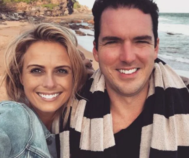 A couple smiling together on a beach, with the ocean and rocks in the background.
