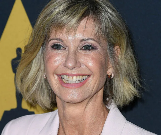 Smiling woman with short blonde hair wearing a light pink blazer against a dark backdrop with a golden statue silhouette.