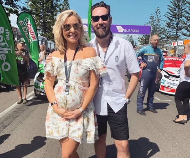 Couple smiling at a motorsport event with race cars and banners in the background.