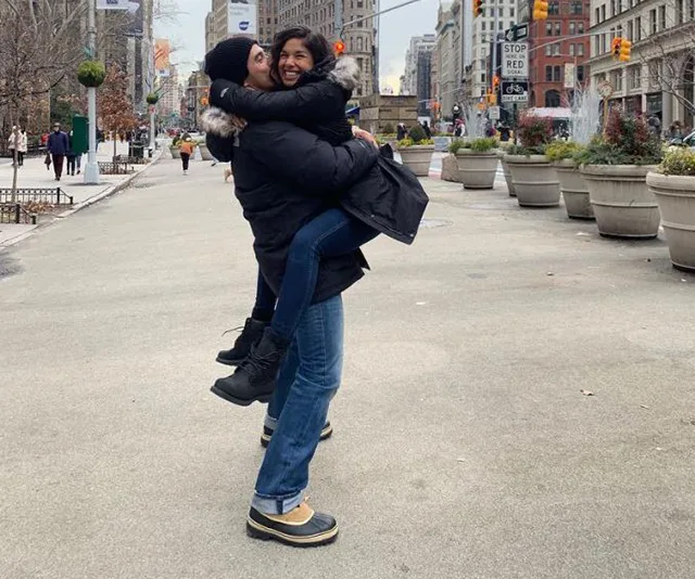 Man in winter clothing lifts and hugs a laughing girl on a city street, surrounded by buildings and potted plants.