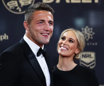 A couple dressed formally smiles on a red carpet at an event with a dark background featuring logos.