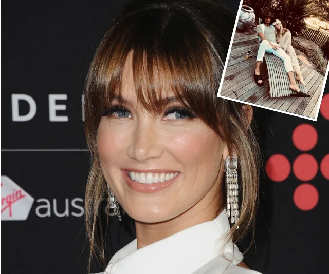 Delta Goodrem smiling at an event with a cutout photo showing her sitting with a man on a wooden bench.