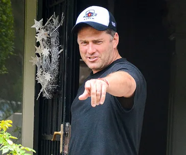 Man in a black t-shirt and cap stands at an open door, pointing forward with a playful expression.