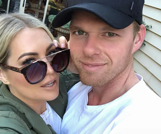 A couple taking a selfie outdoors, smiling at the camera; one wears sunglasses, the other a black cap.