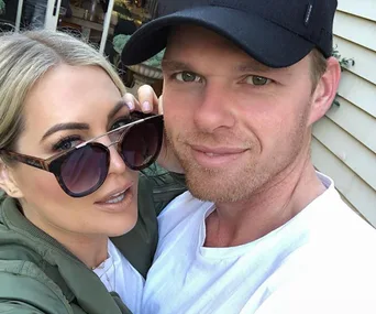 A couple taking a selfie outdoors, smiling at the camera; one wears sunglasses, the other a black cap.