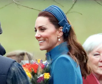 Kate Middleton smiling in a blue coat and headband, holding a bouquet of flowers at a church event.