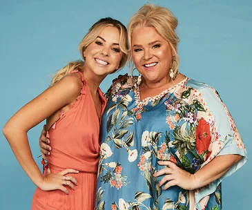 Two smiling women posing together against a plain background, one in a floral dress and the other in a peach dress.