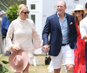 A couple holding hands at an outdoor event, both smiling, with the woman in a lacy dress and man in a blazer and shorts.