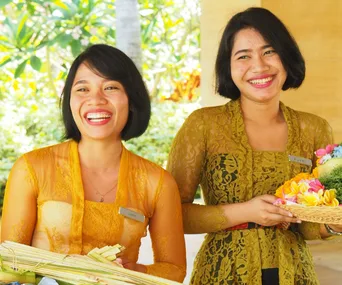 Two smiling women in traditional Balinese attire, holding flower offerings amidst a tropical background.