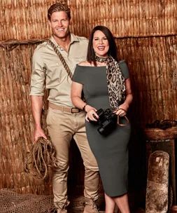 A man and woman in jungle-themed attire, with a rope and camera, in front of a straw backdrop.