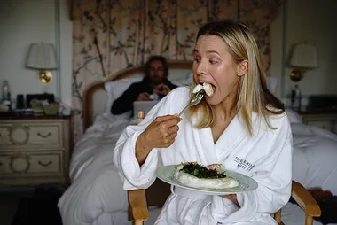 Person in bathrobe eating from a plate in a cozy room, focused on food; another person blurred in background on bed.