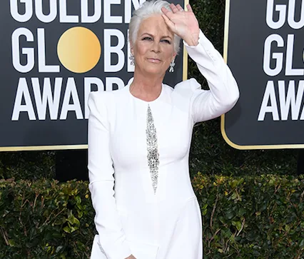 Jamie Lee Curtis in a white dress at the Golden Globe Awards, shielding her eyes with her hand while posing.