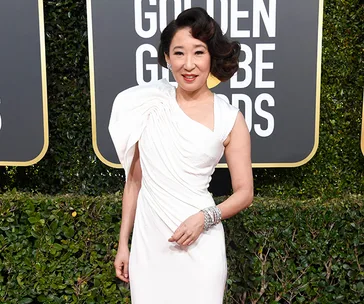 Sandra Oh in a white gown at the Golden Globe Awards, standing in front of a hedge with the event logo in the background.