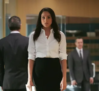 Rachel Zane in an office setting, wearing a white blouse and black skirt, holding documents, with blurred figures behind her.
