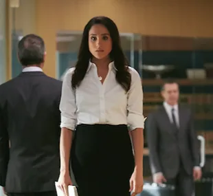 Rachel Zane in an office setting, wearing a white blouse and black skirt, holding documents, with blurred figures behind her.