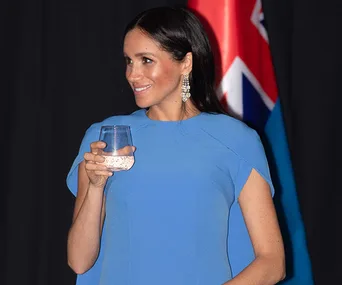 Meghan Markle in a blue dress, holding a glass, wearing diamond earrings, stands in front of a flag.