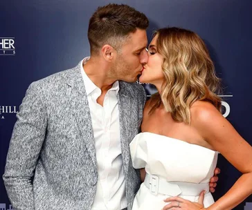 Couple kissing on a red carpet, with the man in a grey blazer and the woman in a strapless white dress.