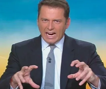 Man in a suit making an animated gesture, on what appears to be a television set with a blurred colorful background.