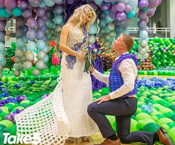 Man proposing to woman in a dress made of white and blue balloons, surrounded by colorful balloon decorations.