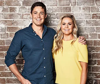 Two people smiling and standing in front of a brick wall, one in a blue shirt and the other in a yellow top.