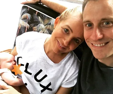 A couple takes a selfie with a baby, sitting on a patterned chair. The woman wears a white shirt.