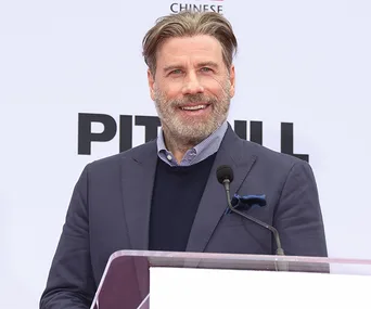 John Travolta smiling at an event, wearing a suit, with text on a white background behind him.