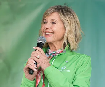 Olivia Newton-John smiling and holding a microphone in a green jacket.