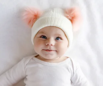 Baby wearing a white hat with pink pom-poms, smiling on a soft white blanket.
