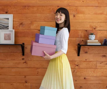 A person holds colorful boxes, smiling in a tidy room with wooden walls and shelves.