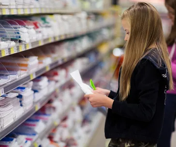 Girl reading a shopping list in a store aisle, surrounded by shelves of school supplies.