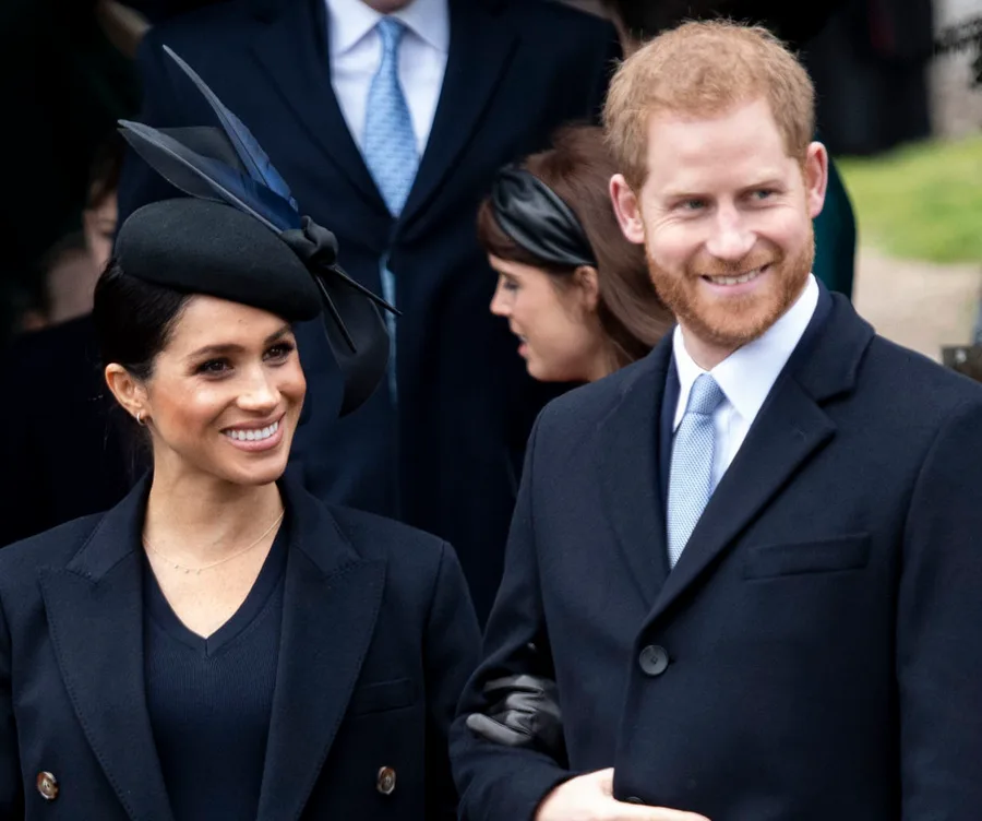 Two individuals smiling, dressed in formal black coats, with a group in the background.