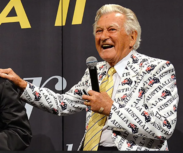 A smiling man in a suit with "Australia" printed on it, holding a microphone at an event.