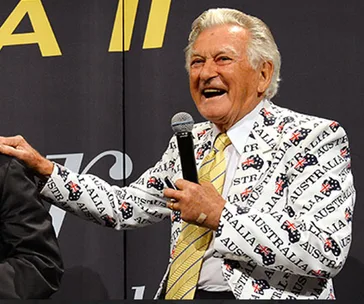 A smiling man in a suit with "Australia" printed on it, holding a microphone at an event.