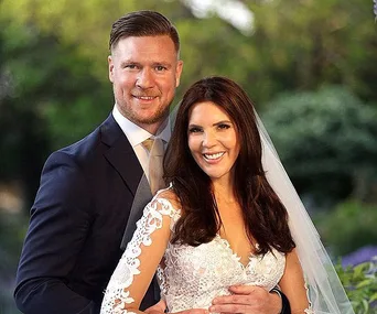 A couple from a wedding show poses, smiling. The bride in lace, groom in a suit, with greenery in the background.