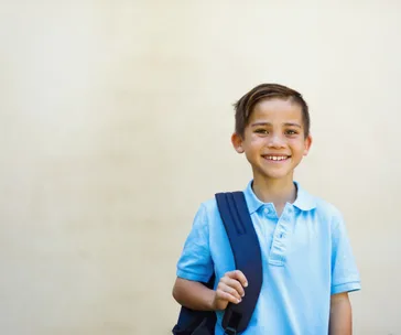 A smiling child in a light blue shirt carrying a dark blue backpack over one shoulder against a plain background.
