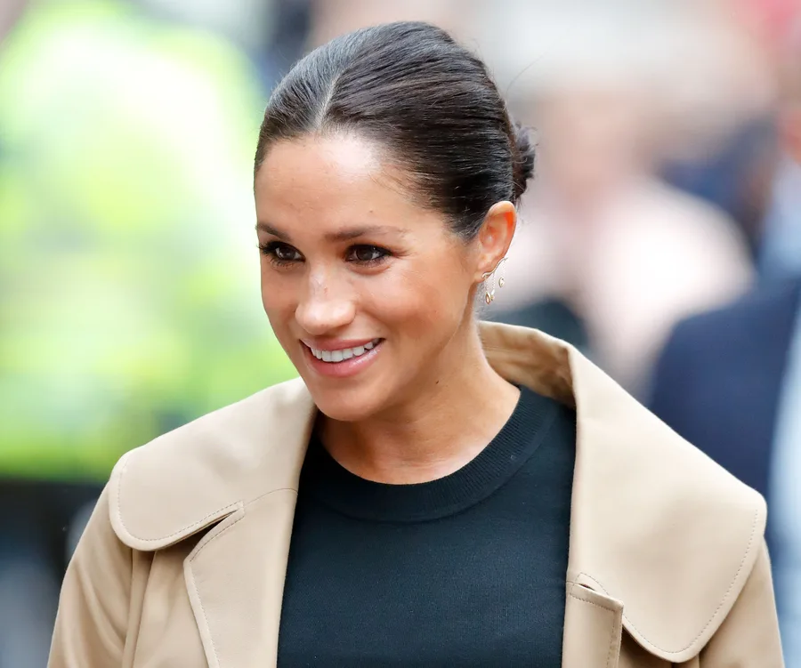 A woman with dark hair tied back, wearing a tan coat and black top, smiling outdoors.