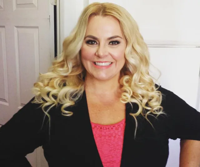 A smiling woman with long curly blonde hair wearing a black top over a pink shirt.