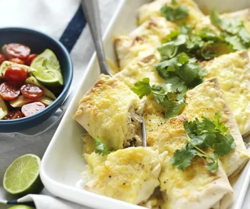 Baked enchiladas topped with cheese and cilantro in a dish; side of cherry tomato salad and lime slices.