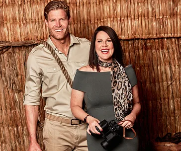 Two people smiling, one holding binoculars, in front of a rustic backdrop, from "I'm a Celebrity...Get Me Out of Here!".