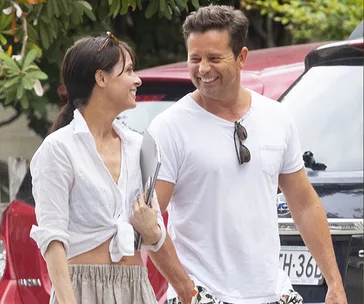 A man and woman smiling at each other while walking outdoors, with cars and trees in the background.