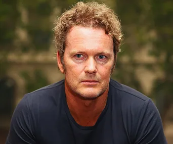 A person with curly hair and a serious expression, wearing a dark shirt, with a blurred background.