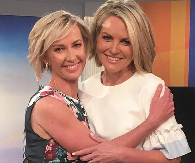 Two women with short blonde hair smiling and embracing each other on a brightly lit set.