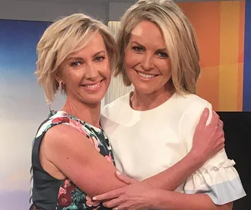 Two women with short blonde hair smiling and embracing each other on a brightly lit set.