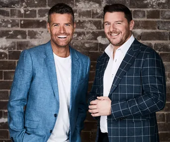 Two men smiling, wearing suits; one in blue plaid, the other in light blue, against a brick background.