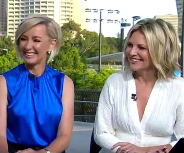 Two women outdoors, smiling during a TV segment, with buildings in the background.