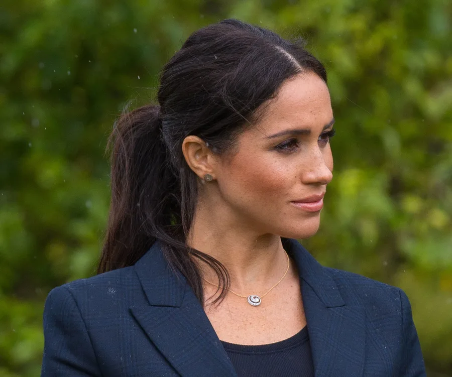 Royal woman with dark hair in a black blazer, looking to the side, set against a blurred green background.