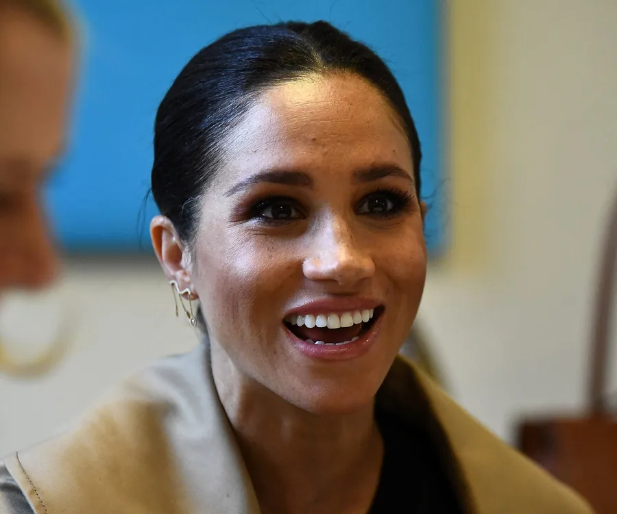 Meghan Markle smiling, wearing earrings and a light-colored coat, with a blue background behind her.